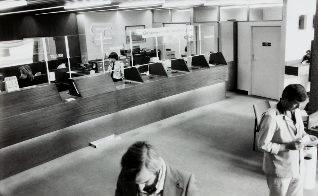 A black and white photo of people waiting at a bank while a teller talks on the phone.